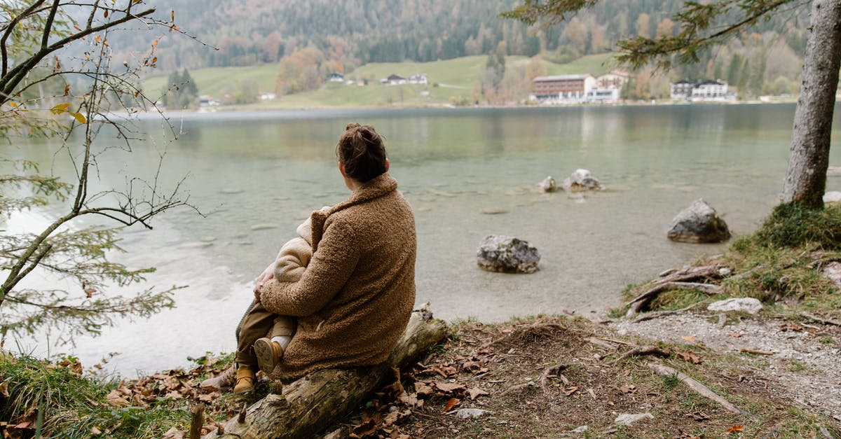 Can a child with accompanied visa travel back to home country with mother - Back view of unrecognizable mother in casual warm outerwear sitting with baby on hands on log near lake and admiring calm scenery in overcast weather Can a child with accompanied visa travel back to home country with mother - Back view of unrecognizable mother in casual warm outerwear sitting with baby on hands on log near lake and admiring calm scenery in overcast weather