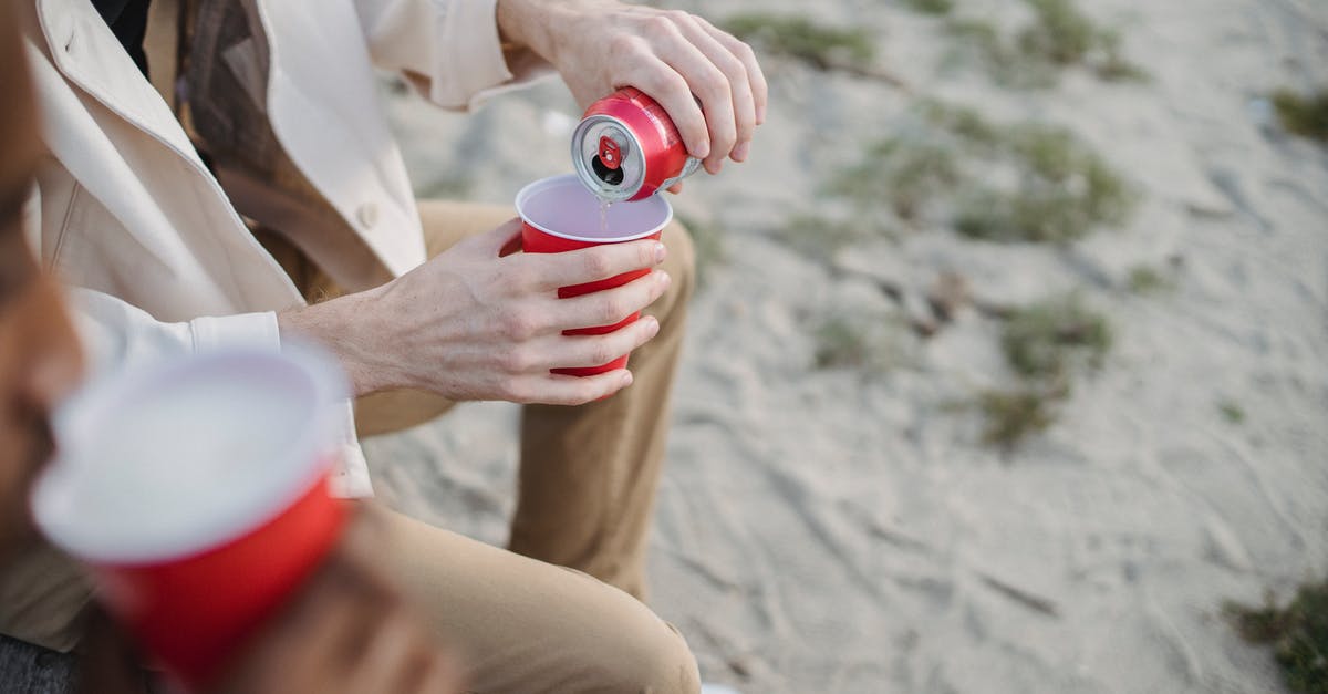 Can anyone identify this beach? - High angle of crop anonymous male pouring refreshing drink into red cup while sitting on sandy shore with black friend