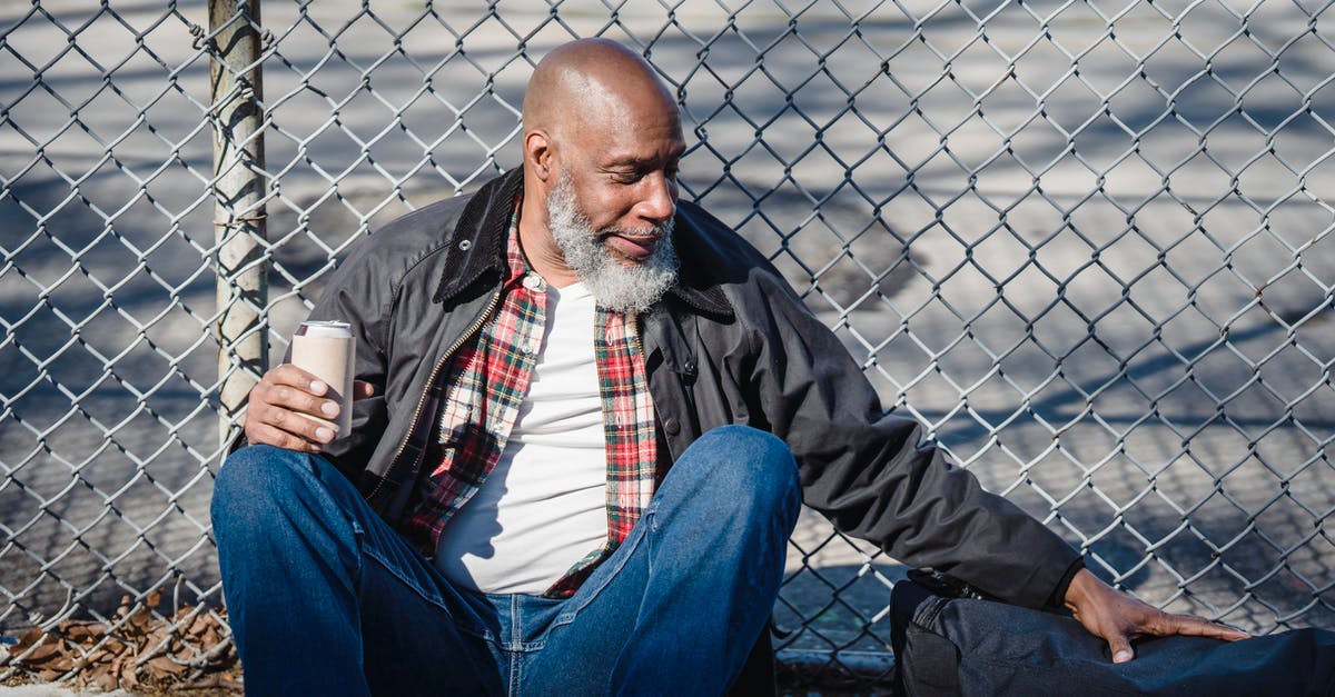 Can growing a beard cause problems in crossing borders? - Side view of black homeless bald man sitting on street with beer can in hand and leaning on wire railing