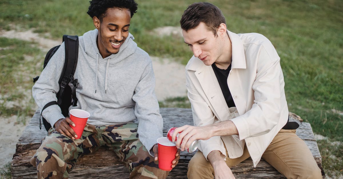 Can I bring duty free liquids through LIM-CDG-LHR? - High angle of male pouring carbonated drink from tin can to smiling black friend sitting with red cup