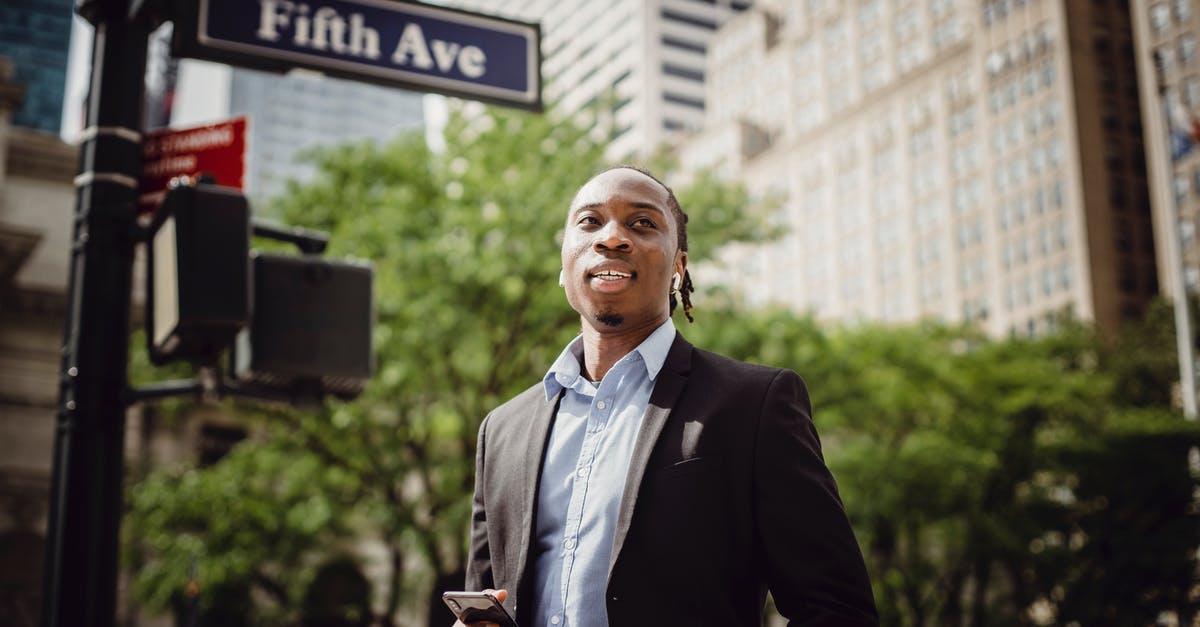 Can I carry 5 mobile phones to US from India? - From below of young handsome black male in smart suit listening to music and waiting for car to pass while standing near street sign in Manhattan on sunny day