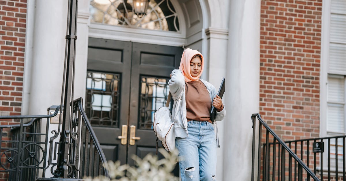 Can I carry a PC in a flight? - Young Muslim woman wearing jeans and orange hijab carrying backpack and laptop walking down stairway after studies in university