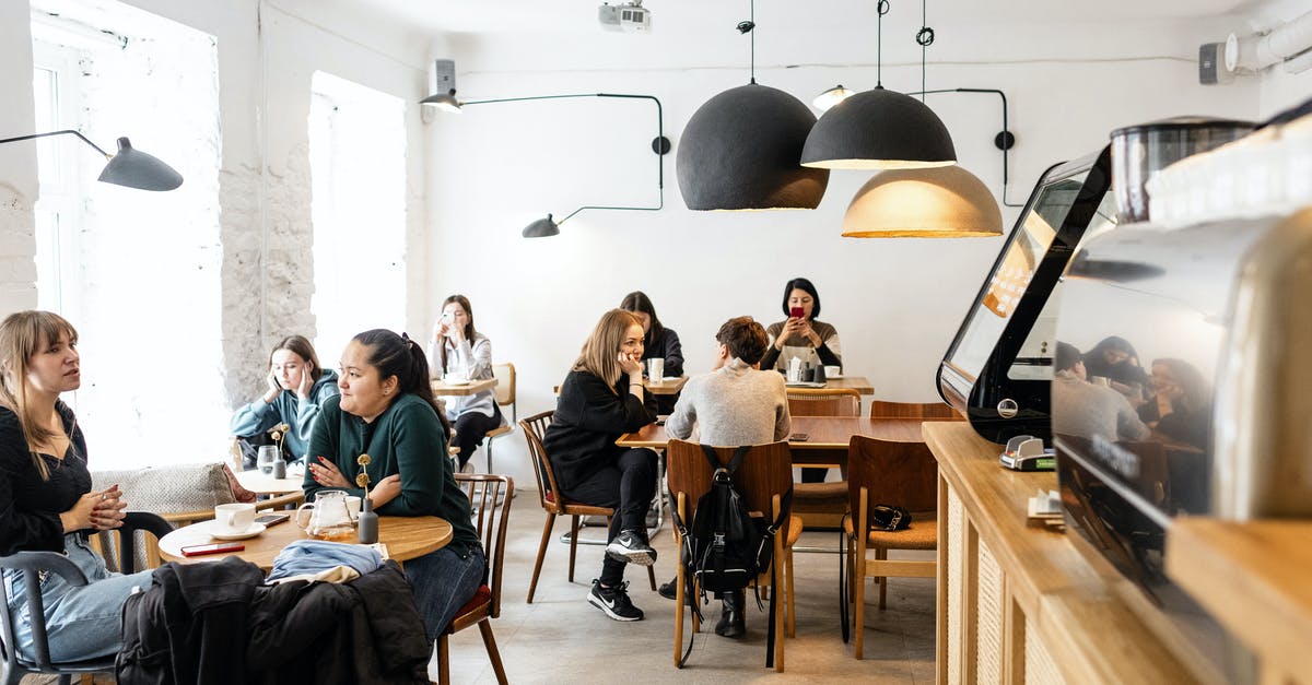 Can I do a multi day cycle tour in Germany now (during Corona time), on my own, camping in public places? - Group of multiracial friends in casual wear sitting at wooden tables in cozy cafeteria near cash box and big lamps while conversing