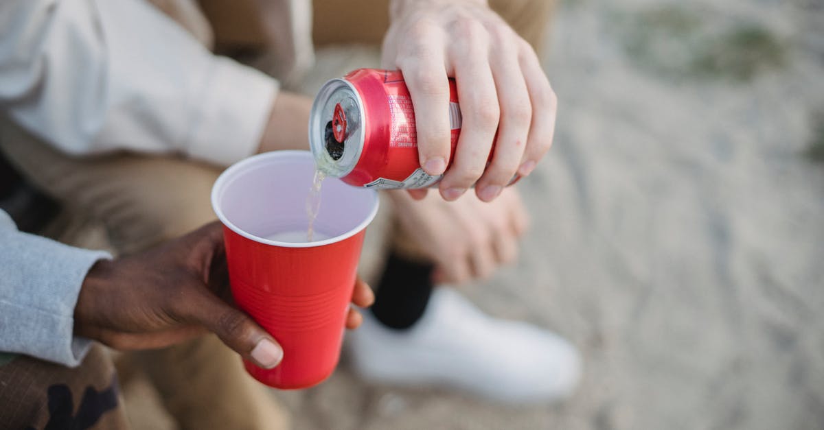 Can I invite a friend to visit me in the UK? - From above of crop anonymous man pouring fizzy drink from can into red plastic cup of black friend while sitting on sandy ground