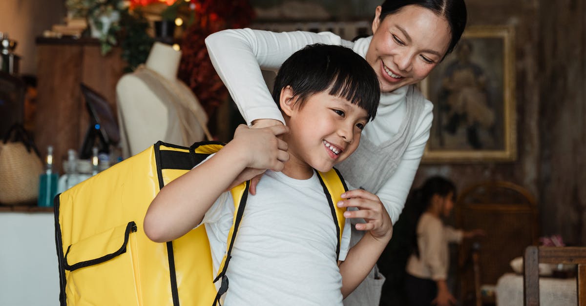 Can I strap a small bag to my backpack so that it still counts as one bag? - Happy female cafeteria employee putting on big yellow thermo bag on shoulders of positive child in casual wear working as food courier and looking away