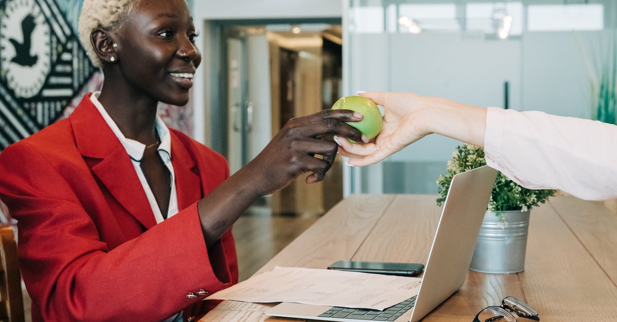 Can I take a laptop into Japan? - Side view of cheerful African American businesswoman in stylish clothes sitting at wooden table with netbook and documents and taking green apple from coworker in modern workspace