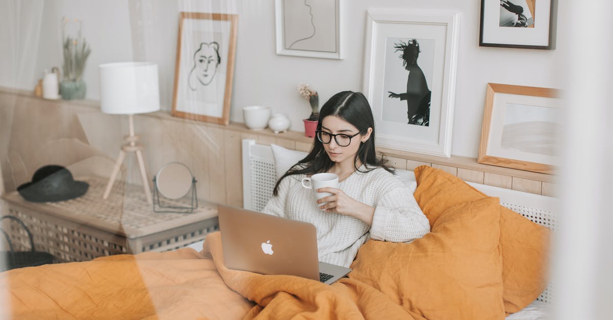 Can I transit through UK using Business Visitor Visa? - Focused young brunette in eyeglasses with cup of hot drink lying in comfortable bed and working on laptop in morning