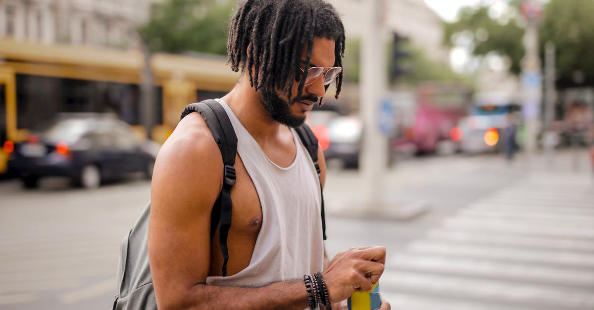 Can i travel to Spain? - Confident adult Hispanic male traveler in sunglasses and casual clothes with backpack opening can of drink while walking along pavement against blurred cars driving along street and empty crosswalk in downtown