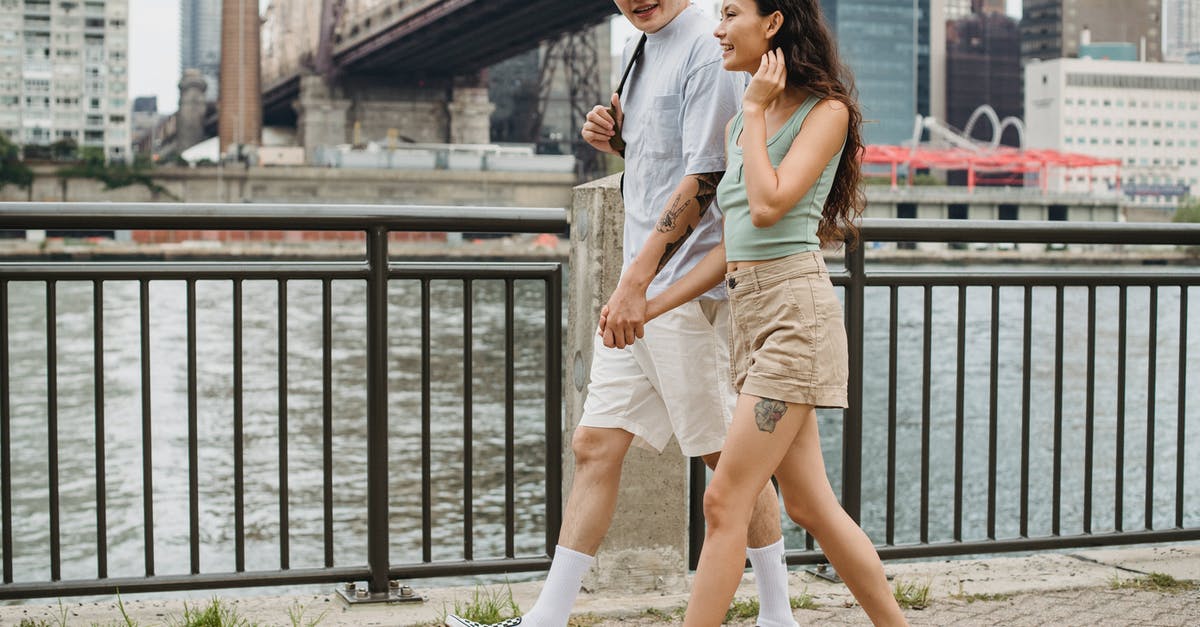 Can I travel to the US without holding a job? - Stylish diverse couple holding hands and strolling on city promenade under bridge over river