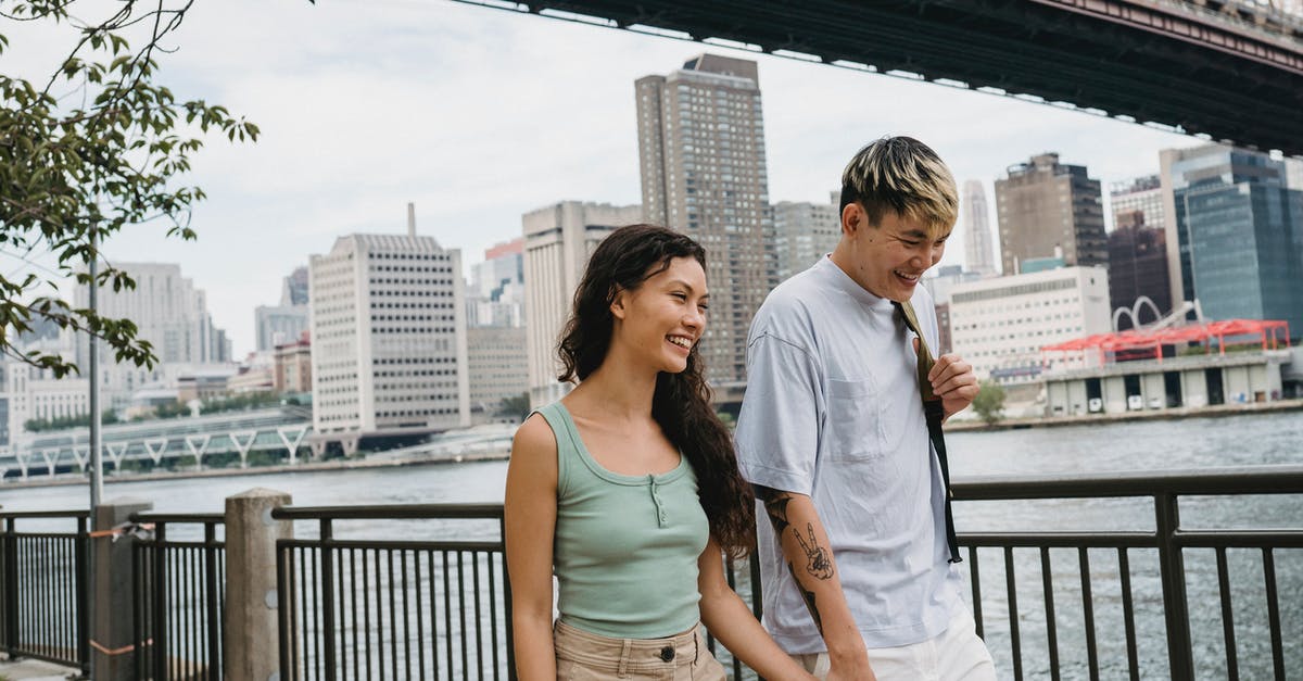 Can I travel to the US without holding a job? - Positive young Asian man and ethnic woman holding hands and smiling while walking together on quay near bridge over river in New York