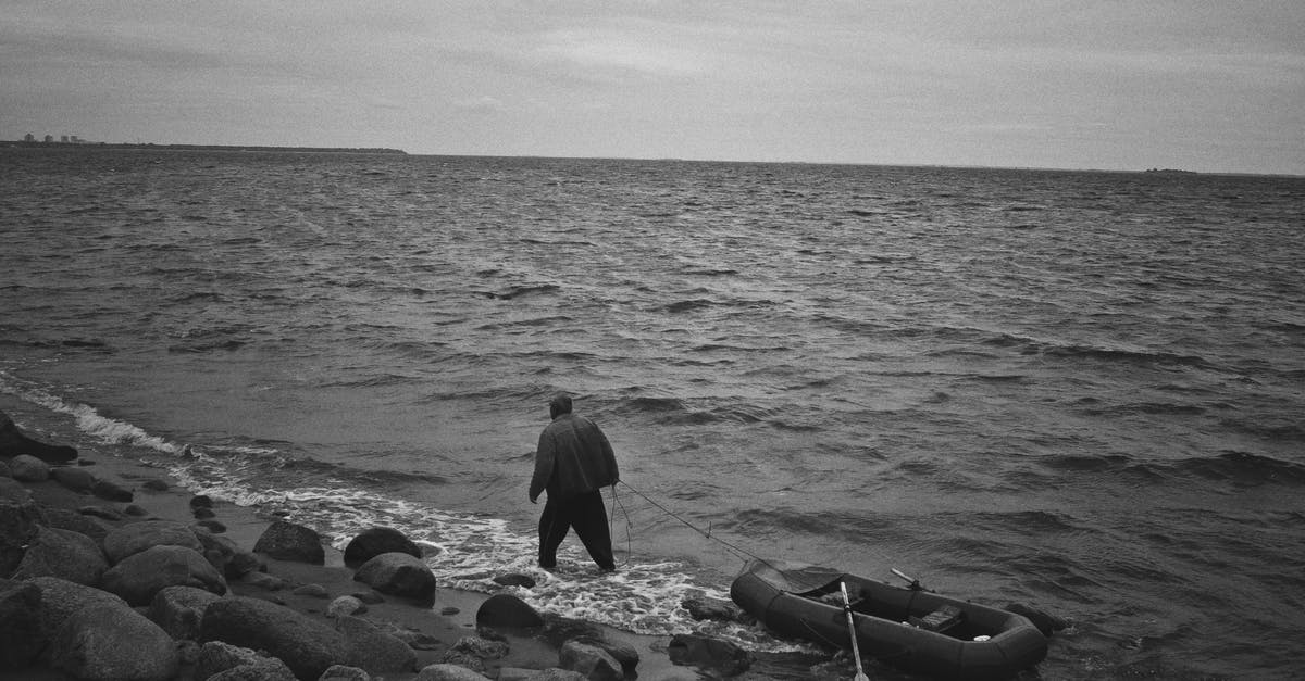 Can I walk around Easter Island alone at night? - From above black and white of full body anonymous male in black outfit with inflatable boat walking along coast of sea with stones