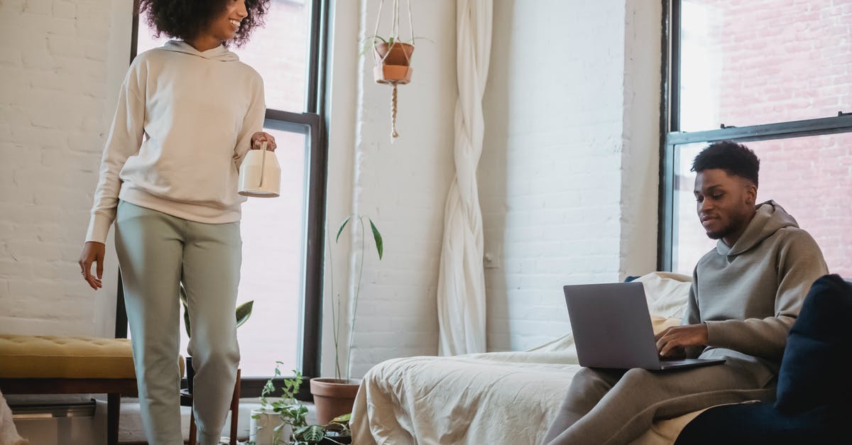 Can one walk to the Patriarchate of Peć? - Happy young black woman with Afro hair in casual outfit smiling and walking in living room with watering can in hand near boyfriend working remotely on laptop