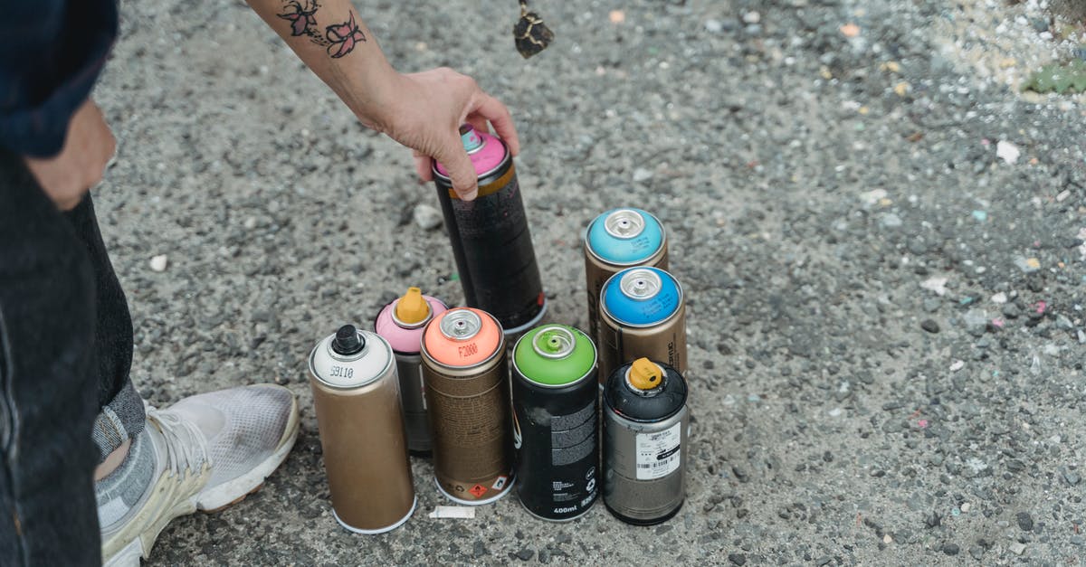 Can you identify this city or country? [closed] - Crop anonymous person in sneakers with tattoo and heap of multicolored spray paint cans on ground standing on street in city