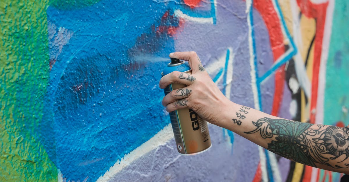 Can you identify this city or country? [closed] - Crop unrecognizable tattooed painter spraying blue paint from can on multicolored wall with creative graffiti while standing on street in city