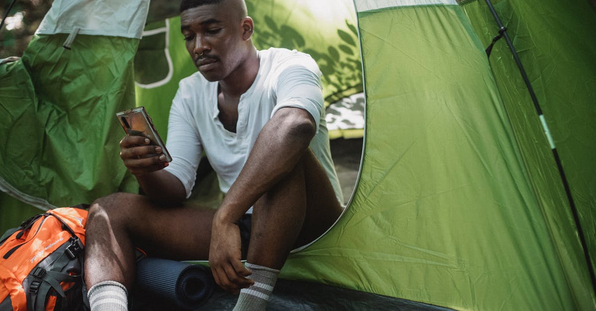 Canada tourist visa online appication - Sad African American male traveler resting in tent and browsing Internet on cellphone while spending journey in nature