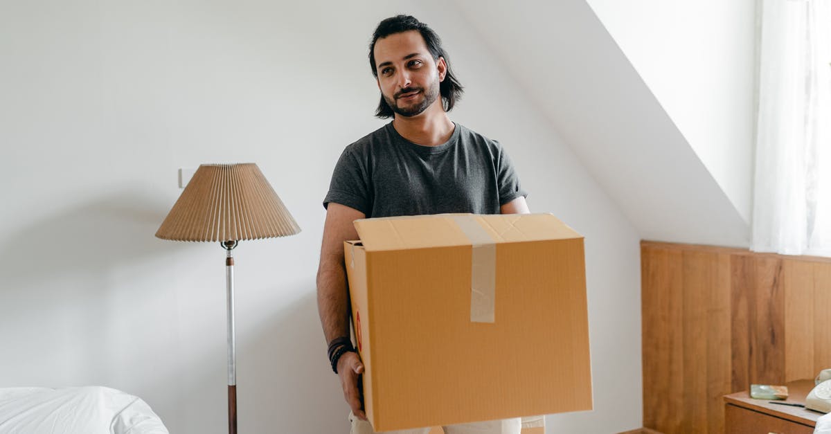 Canadian Temporary Resident Visa fee for passport delivery - Cheerful young bearded male in casual wear carrying cardboard container in sunny bedroom while moving into new apartment