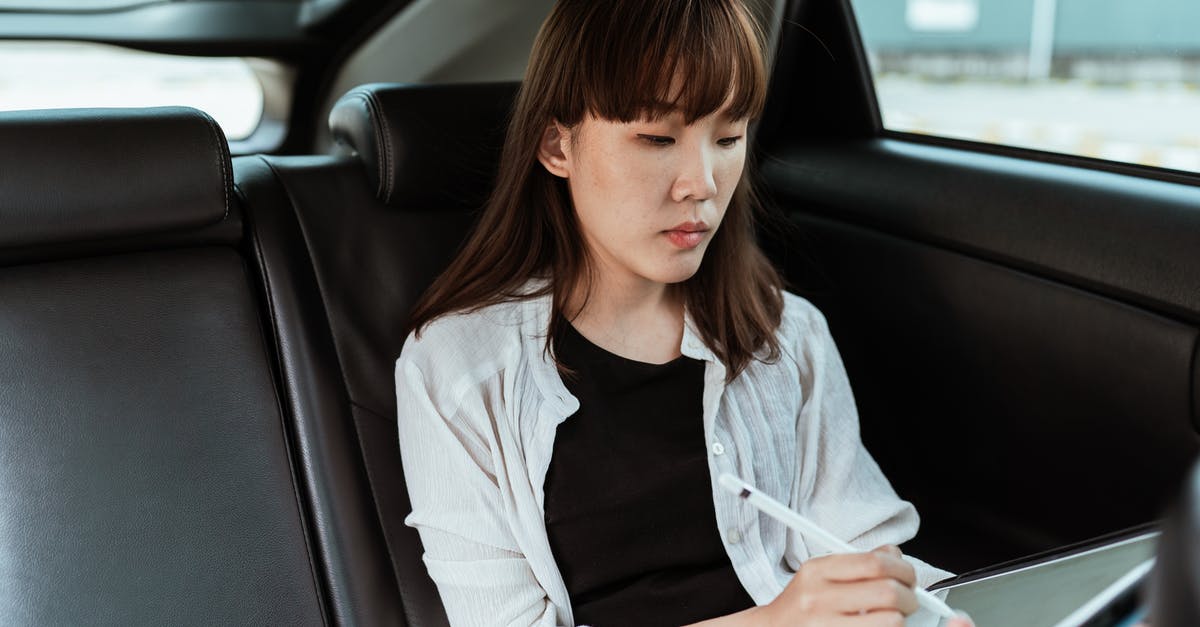 Cancelling Schengen Student Visa Application - Concentrated Asian female in casual clothes working on modern tablet with stylus while riding in car backseat