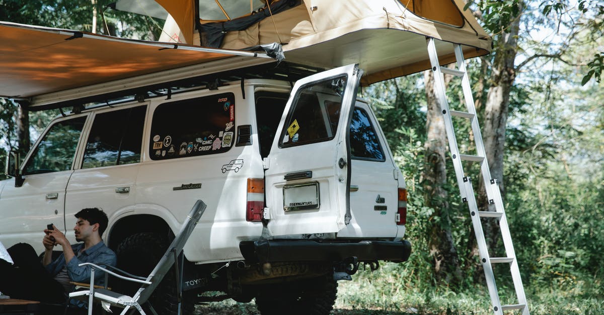 Car camping on transatlantic ship - Man sitting under awning placed near offroader with tent on roof in forest