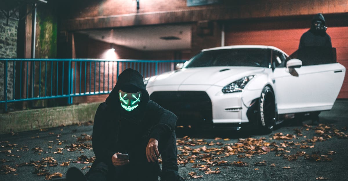 Car mask rules in Italy when living together during holiday - Anonymous male in neon mask browsing internet on cellphone while sitting on asphalt road covered with faded leaves near modern auto and friend standing on illuminated street at night