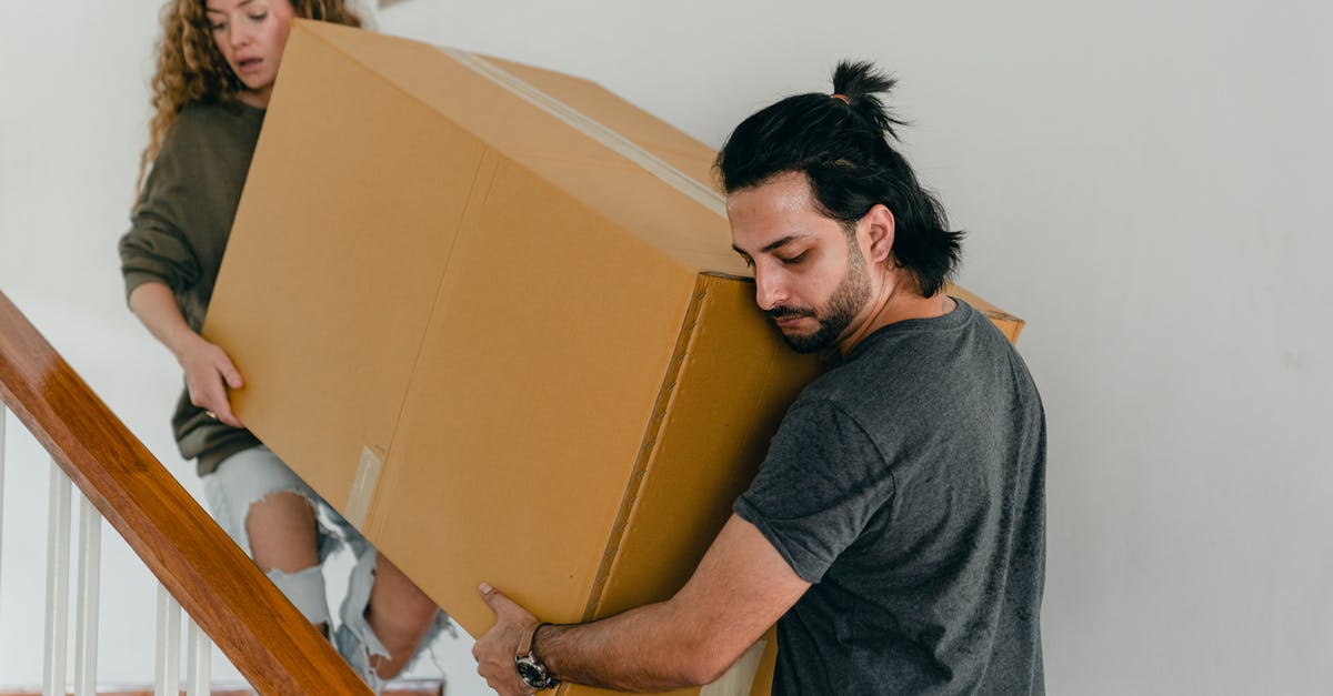 Carry bicycle into the US as personal item - Focused couple in casual clothes carrying big carton box together down stairs in new house while moving personal items