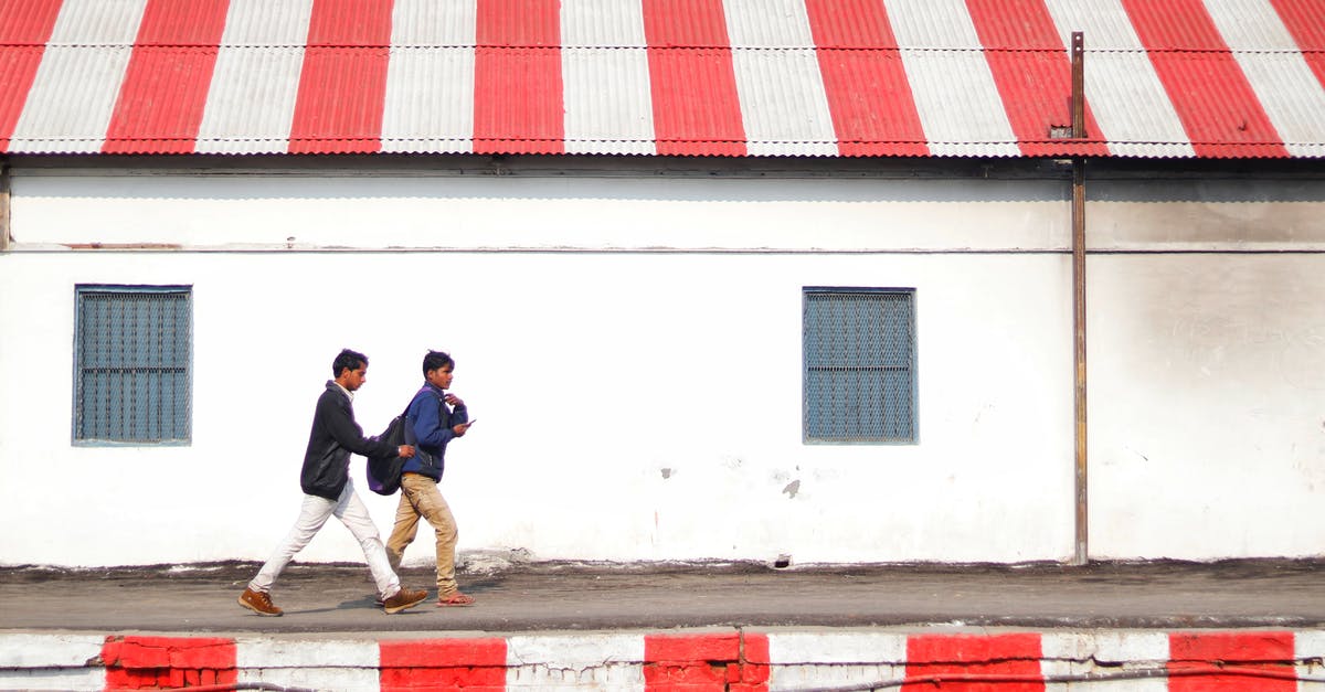 Change of boarding point on Indian Railways [duplicate] - Side view of young Asian guys walking on platform painted with white and red stripes along shabby stone building with striped white and red roof on daytime