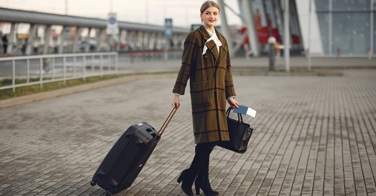 Changing baggage final destination - hidden-city ticketing - Full body young female passenger in trendy plaid coat with suitcase strolling on pavement near modern building of ariport terminal taking bag and documents