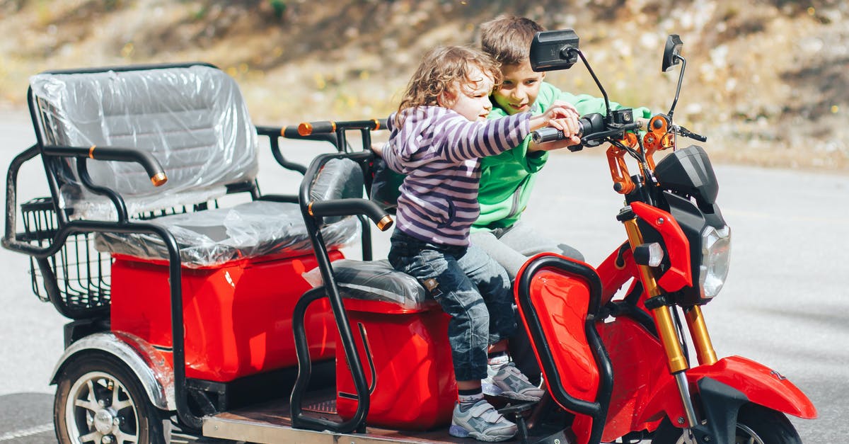 Cheap and convenient way to travel with kids around US - Adorable child helping little brother to ride modern red trike motorcycle on asphalt road on sunny day