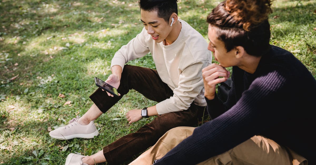 Cheap phone calls from Canada to Poland - From above content young ethnic male friends wearing casual outfits surfing internet on modern mobile phone and sitting on grass in sunny park