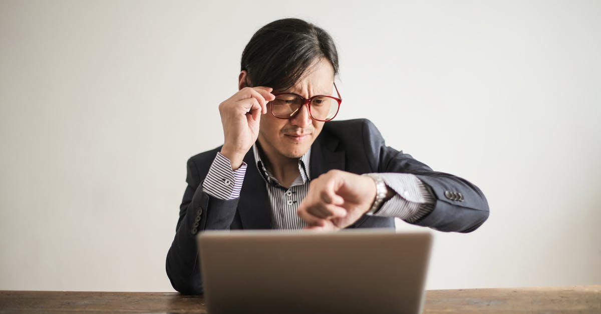 Check in time for layover at Sydney airport - Young frowning man in suit and glasses looking at wristwatch while waiting for appointment sitting at desk with laptop