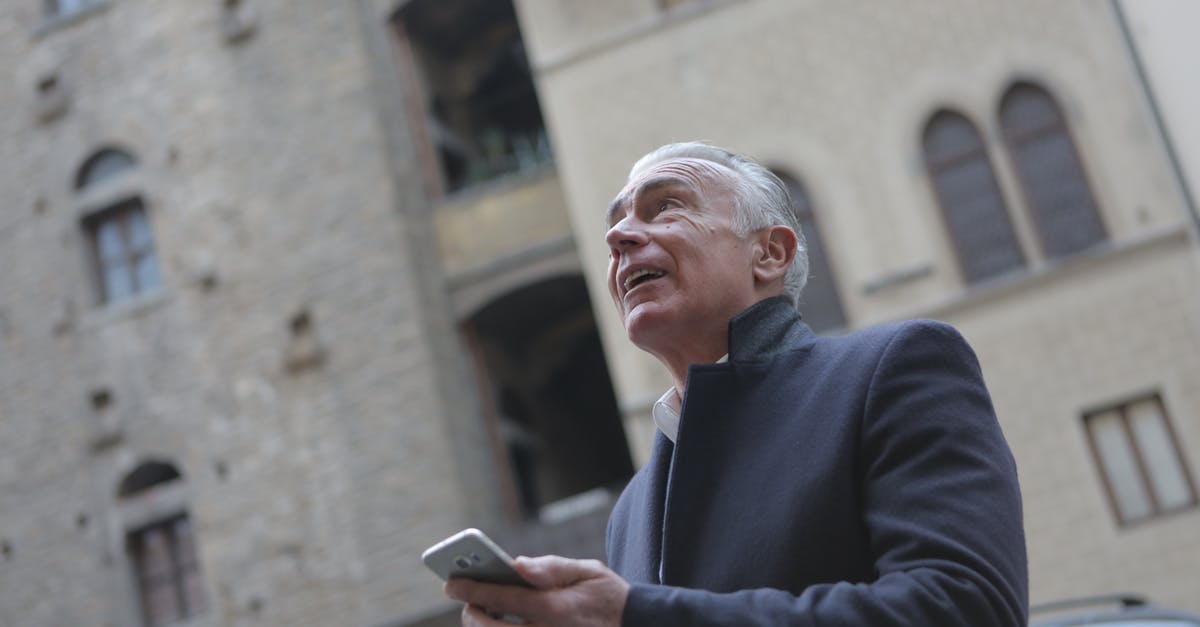 Check in to 2 hotels at same location - From below of mature gentleman in classy wear standing near building in city and checking message on cellphone while looking up