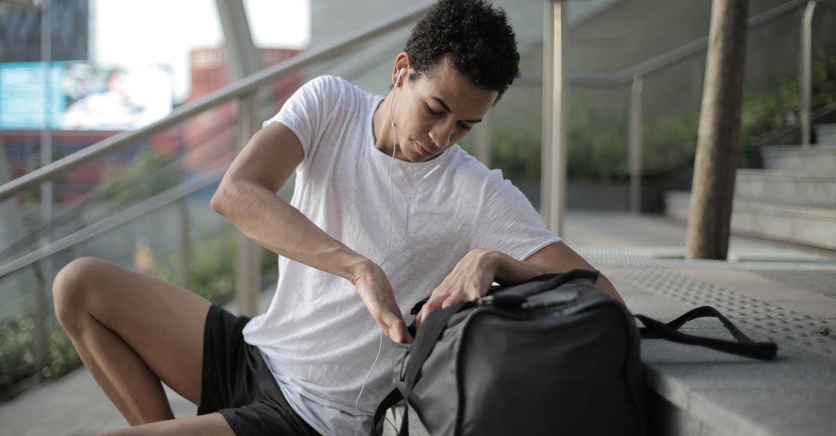 Check-in Luggage - Serious thoughtful African American man in earphones and sports clothes searching in bag pocket device while sitting on stairs and listening to music at entrance of contemporary building in downtown