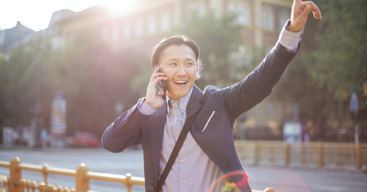 China intercity taxi / taxi-like transport - Happy adult ethnic man in jacket waving with hand hailing taxi on sunny street while speaking on smartphone