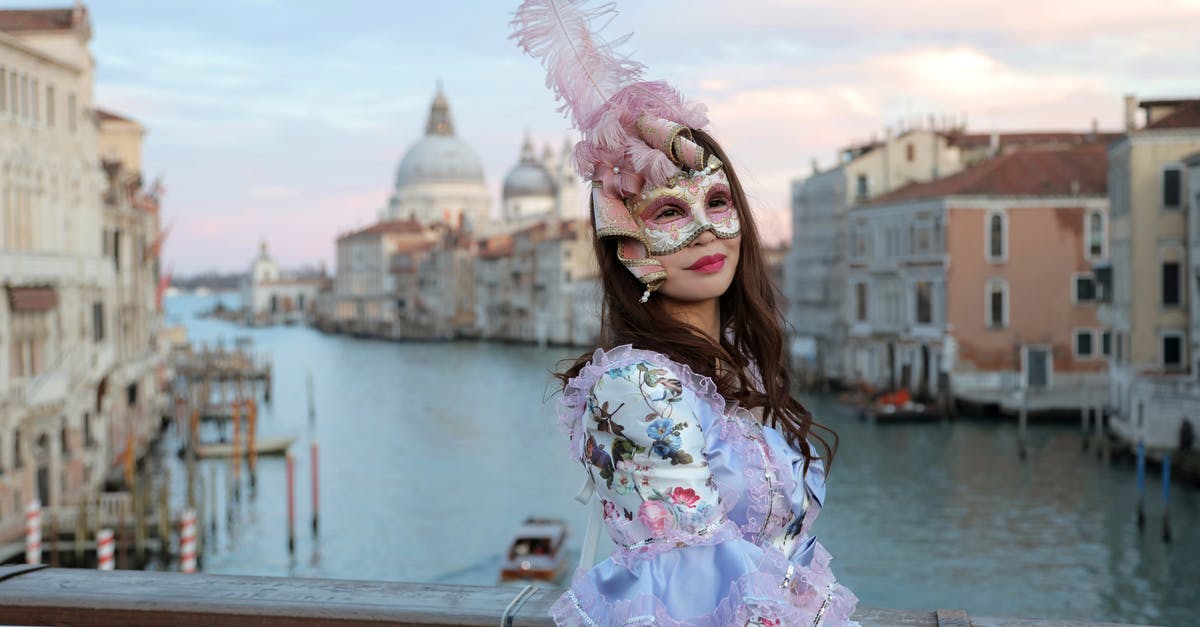 Choosing the Airline for a long-haul fligt, Europe to Taiwan - Side view of unrecognizable woman in magnificent costume and gorgeous Venetian mask with feathers standing on bridge in middle of Grand canal against background of cathedral of Santa Maria della Salute in Venice in Italy and looking at camera