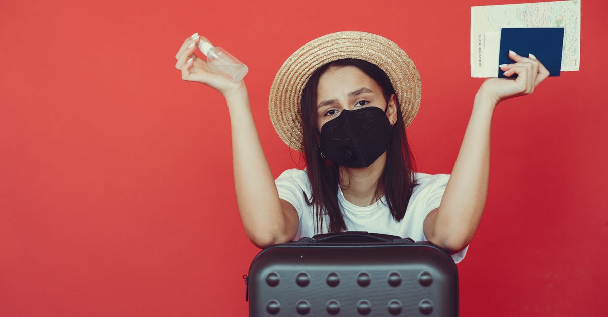 Collecting baggage when transiting & quarantine rules - Young woman in medical mask and wicker hat holding passport and sanitizing spray while sitting behind suitcase and shrugging hand on red background