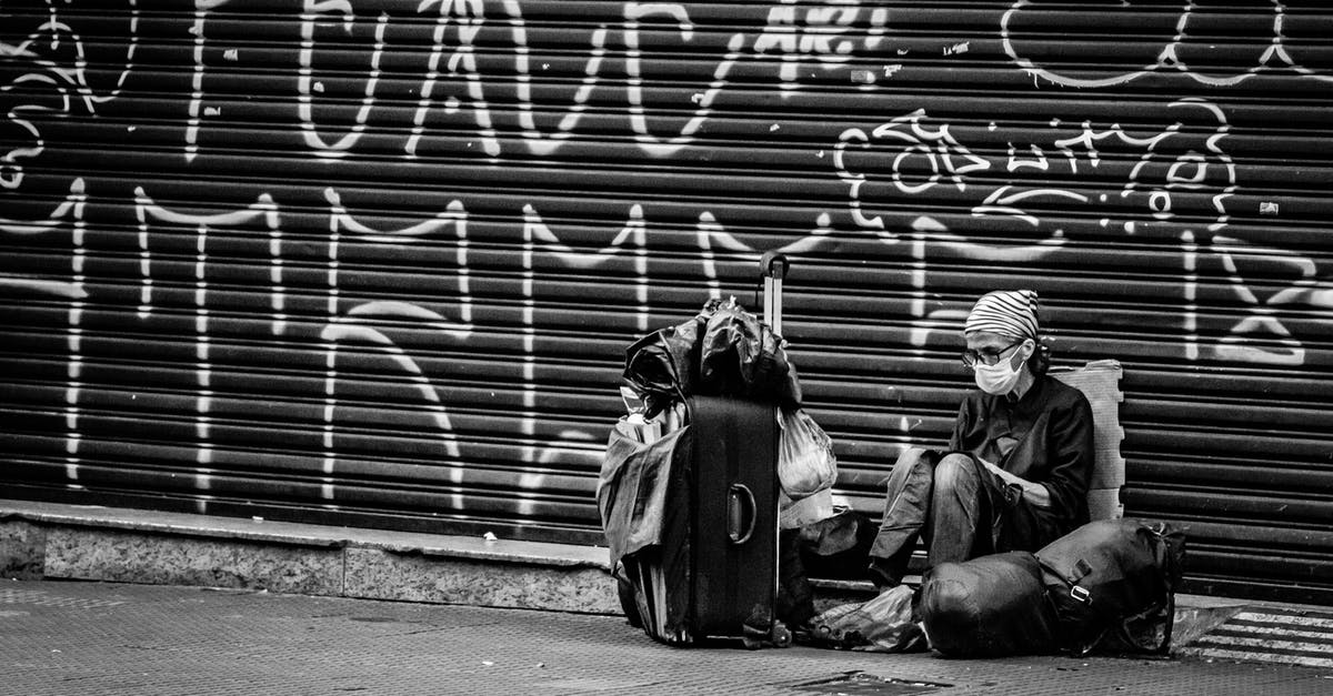 Collecting baggage when transiting & quarantine rules - Black and white of anonymous traveler in sterile mask and eyewear sitting on road near ribbed graffiti wall and backpack during COVID 19 pandemic