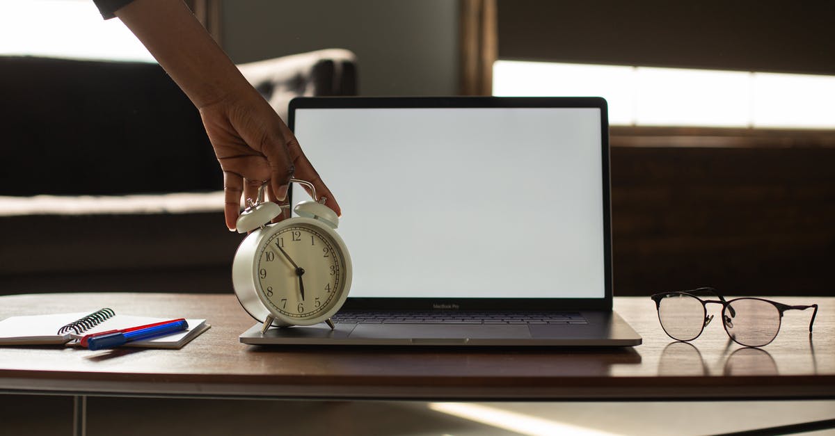 Connecting time ay JFK - Anonymous black person touching alarm clock near opened netbook with white blank screen placed on table with notebook and pens