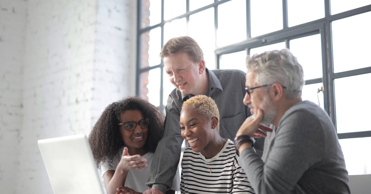 Connection flights with different airlines - Positive diverse coworkers of different ages sitting at table and browsing laptop while discussing project in modern workspace with big window