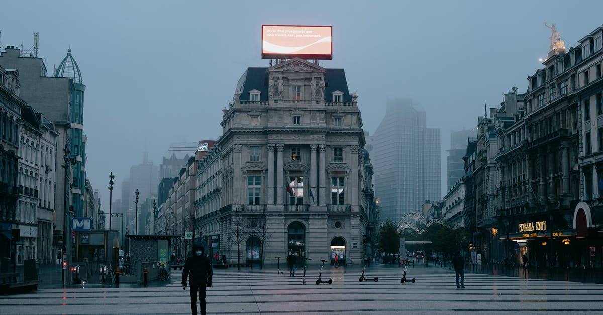 Coronavirus and flight cancellations in Europe - Unrecognizable pedestrians in face masks walking on city square near aged buildings against foggy sky in evening in Brussels