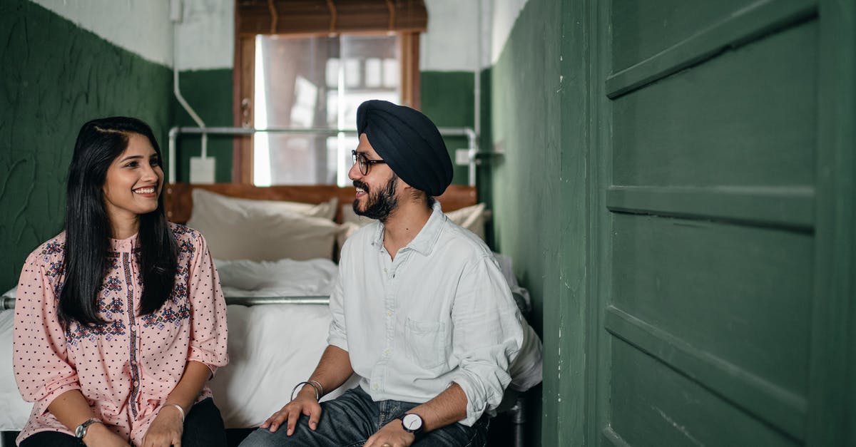 Date of arrival on an Indian e-visa - Smiling Indian couple in casual clothes and turban sitting on bench against bed in narrow green bedroom and chatting while looking at each other happily