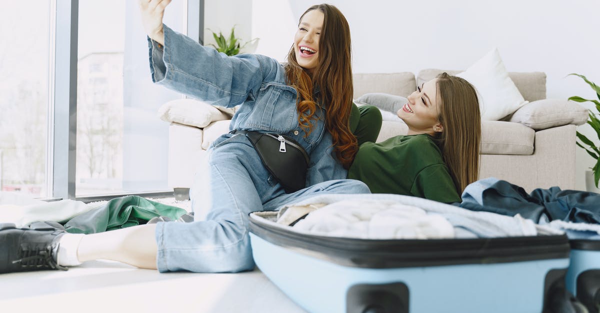 Do I have to recheck my luggage in Chicago? - Happy young female friends in trendy outfits having fun and taking selfie while packing suitcase in cozy room