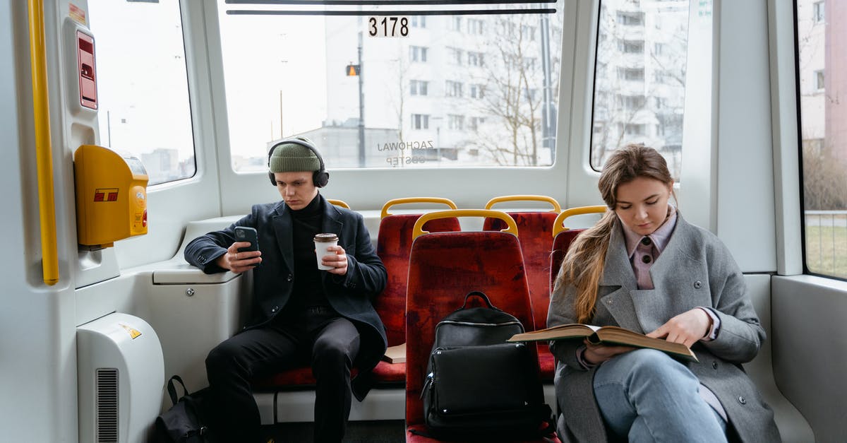 Do I need a transit visa for a Russian overflight? - A Woman in Gray Coat Reading a Book while Sitting Near the Man Wearing Headphones while Holding His Mobile Phone