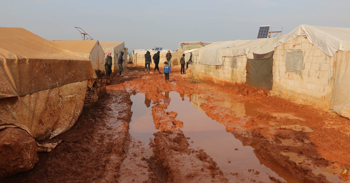 Do I need a visa to walk over the Rainbow bridge? - Group of distant adults and kids strolling on wet dirty ground with puddles near worn out tents in poor area