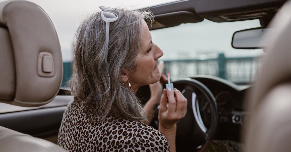 Do North-American car rental agencies typically apply a minimum driving license duration? - Side view of middle aged female with gray hair in stylish outfit applying lip gloss while driving modern cabriolet