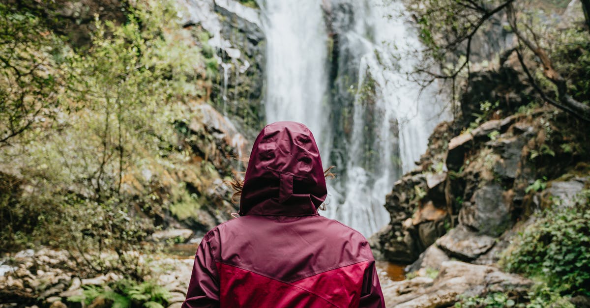 Do the Canadian Rockies lack red foliage in autumn? - Back view of anonymous tourist in red outerwear with hood standing near flowing stream of water falling from rocky cliff in daylight