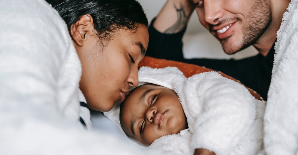 Does changing latitude affect sleep? - High angle of crop young African American woman kissing sleeping cute newborn baby while resting in bed with happy husband