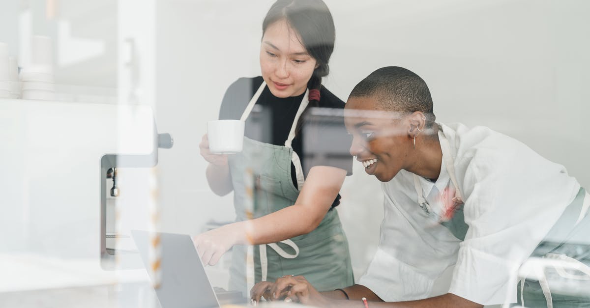 Does StarAlliance's loyality program have a "recommend a friend" program? - Young inexperienced African American female worker smiling and looking at laptop screen and Asian head barista explaining how special program works both shot through glass