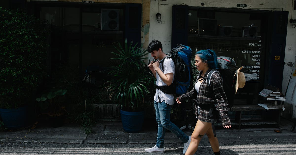 Does the free ASEZA visa allow to travel outside Aqaba? - Side view of young diverse couple of tourists in casual clothes and with backpacks strolling together along city street near shabby stone building with potted plants