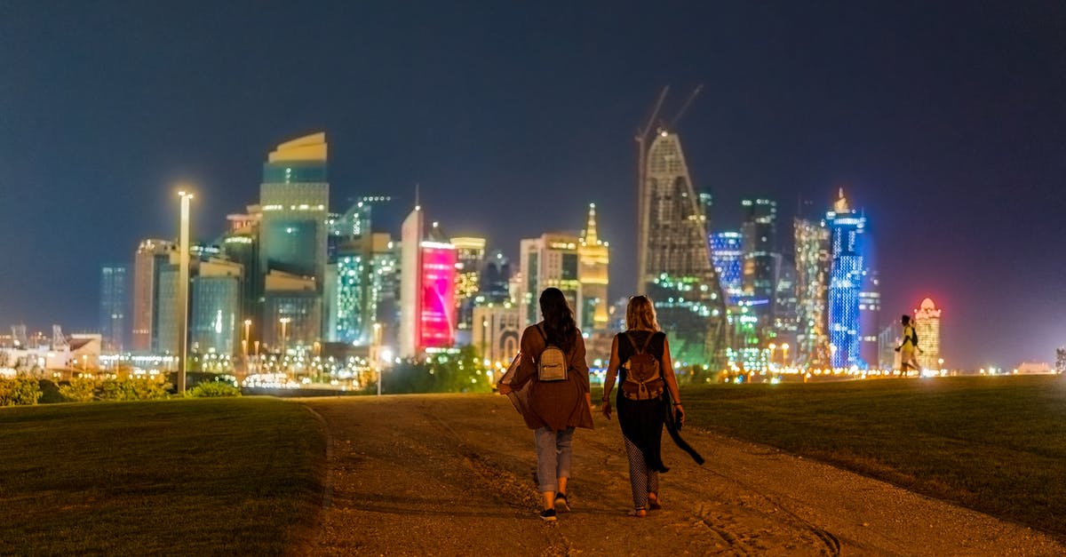 Doha layover at night - Back view of unrecognizable female tourists in casual clothes and backpack walking on sandy road towards modern illuminated buildings in Doha at night