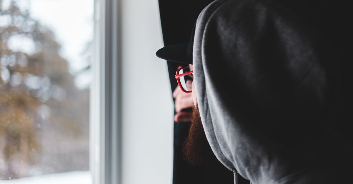 Domestic Or International Check In - Side view of bearded man in glasses and hoodie pulling curtain and looking out window on winter day at home