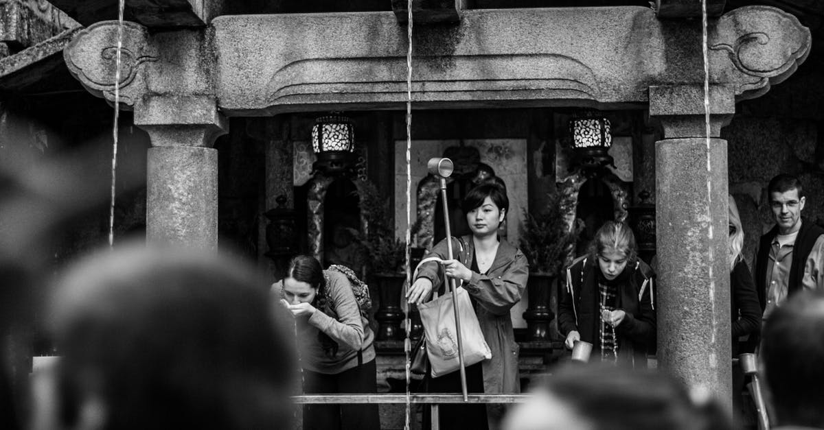 Drinking from the Chalice Well in Glastonbury (Sacred Spring in England) - Black and white of multiracial tourists drinking holy water while standing near old stone construction with narrow vertical pouring flows in sacred place